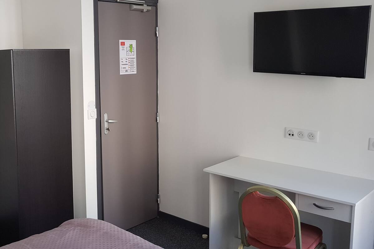 Quarto de hotel com cama, guarda-roupa, pia, secretária e televisão.