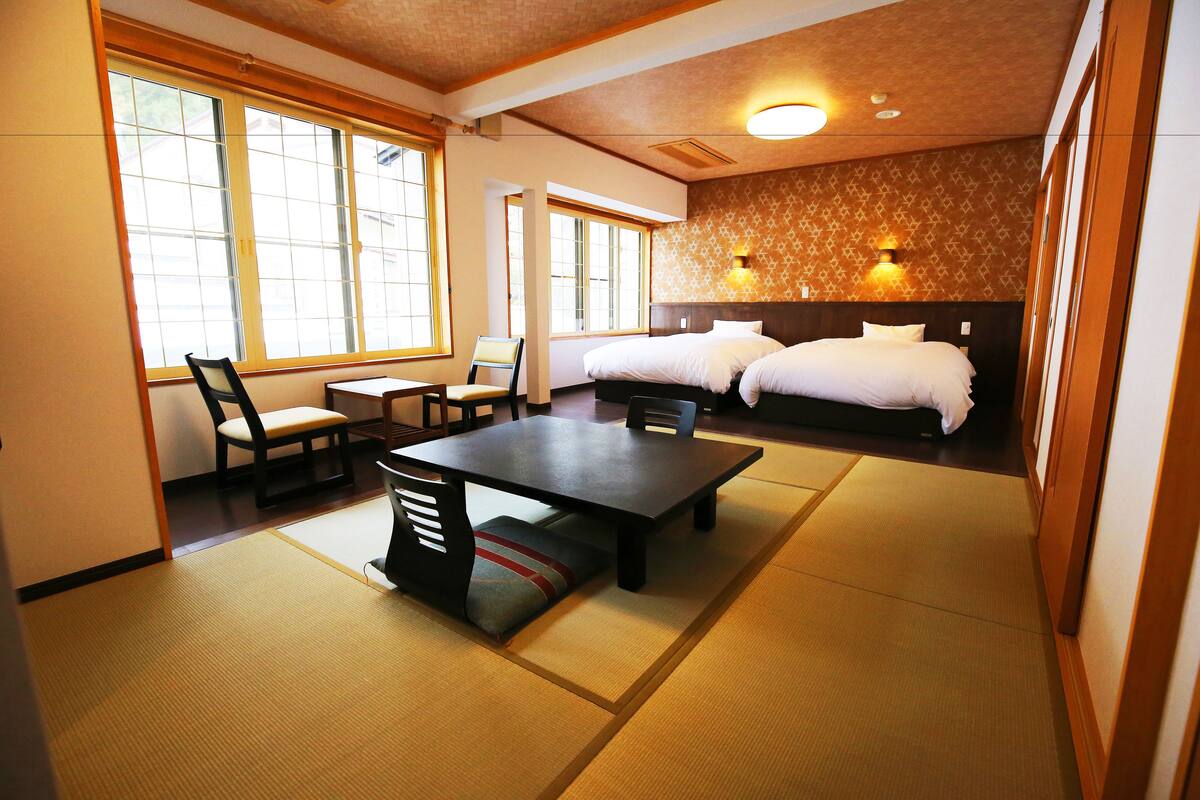 Habitación de hotel con suelo de tatami, una cama de estilo japonés tradicional, una mesa de comedor con sillas y ventanas amplias que ofrecen luz natural.