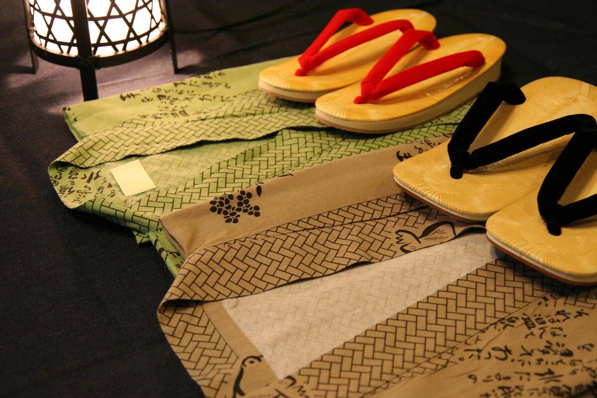 Una mesa con varios objetos, incluyendo un par de sandalias amarillas con correas negras, un par de sandalias rojas con correas negras, y varias esterillas dobladas con diseños tejidos y caracteres japoneses.