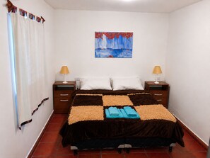 A neatly made bed with a checkered bedspread, two bedside lamps, a painting on the wall, and a curtain on the window.