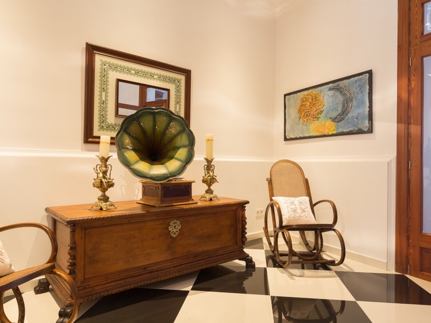 Um quarto com piso xadrez, um armário de madeira com castiçais de latão, um gramofone, uma cadeira de vime e duas obras de arte emolduradas na parede.