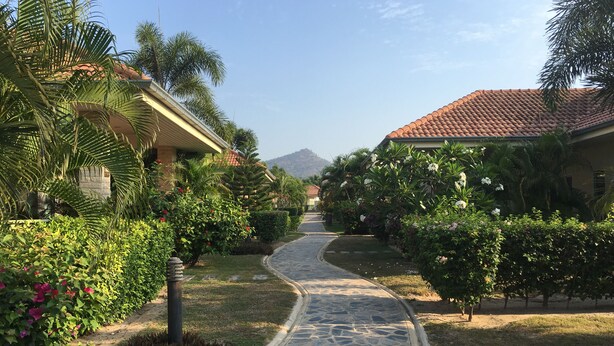 A paved pathway leading through a well-maintained garden with various plants and trees, flanked by buildings with red-tiled roofs. - Nice Villa in Hua Hin (Hua Hin)