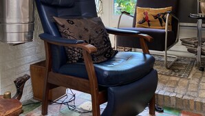 Une pièce comprenant un fauteuil en cuir bleu, une chaise en bois avec un coussin à motifs, une table en métal et une vue sur la verdure à l’extérieur.