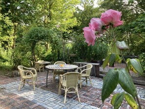 A patio with wicker furniture and a brick floor, surrounded by lush greenery and trees. - 5 pers - Equipped house Gudrun with sauna - Dogs are welcome (Anjum)