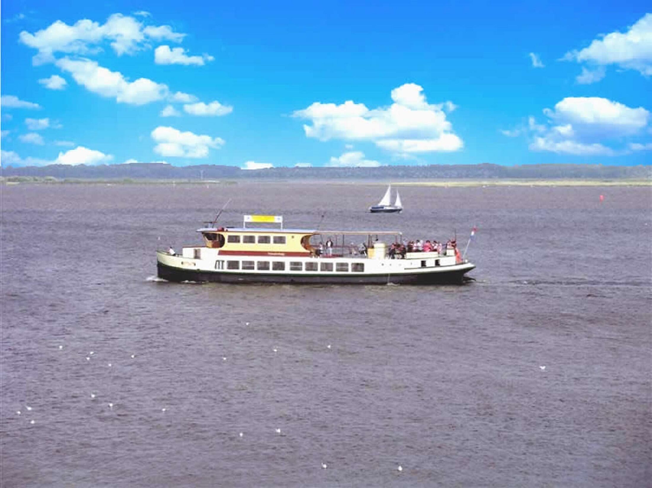 Eine Fähre mit mehreren Decks und Passagieren fährt auf ruhigem Wasser, im Hintergrund ein Segelboot und ein klarer Himmel.