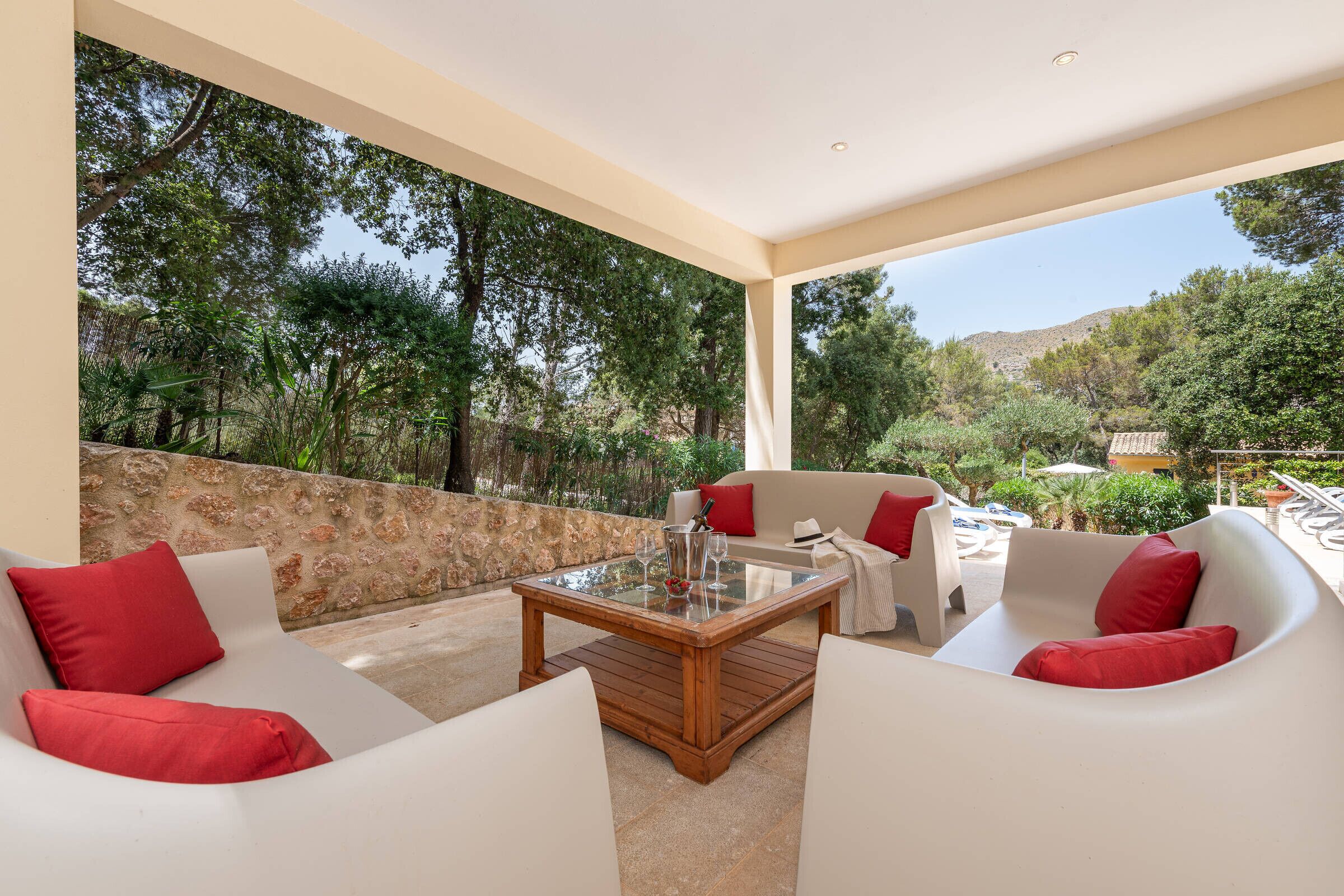 A covered outdoor seating area with white furniture and red cushions, overlooking a natural landscape with trees and a stone wall.