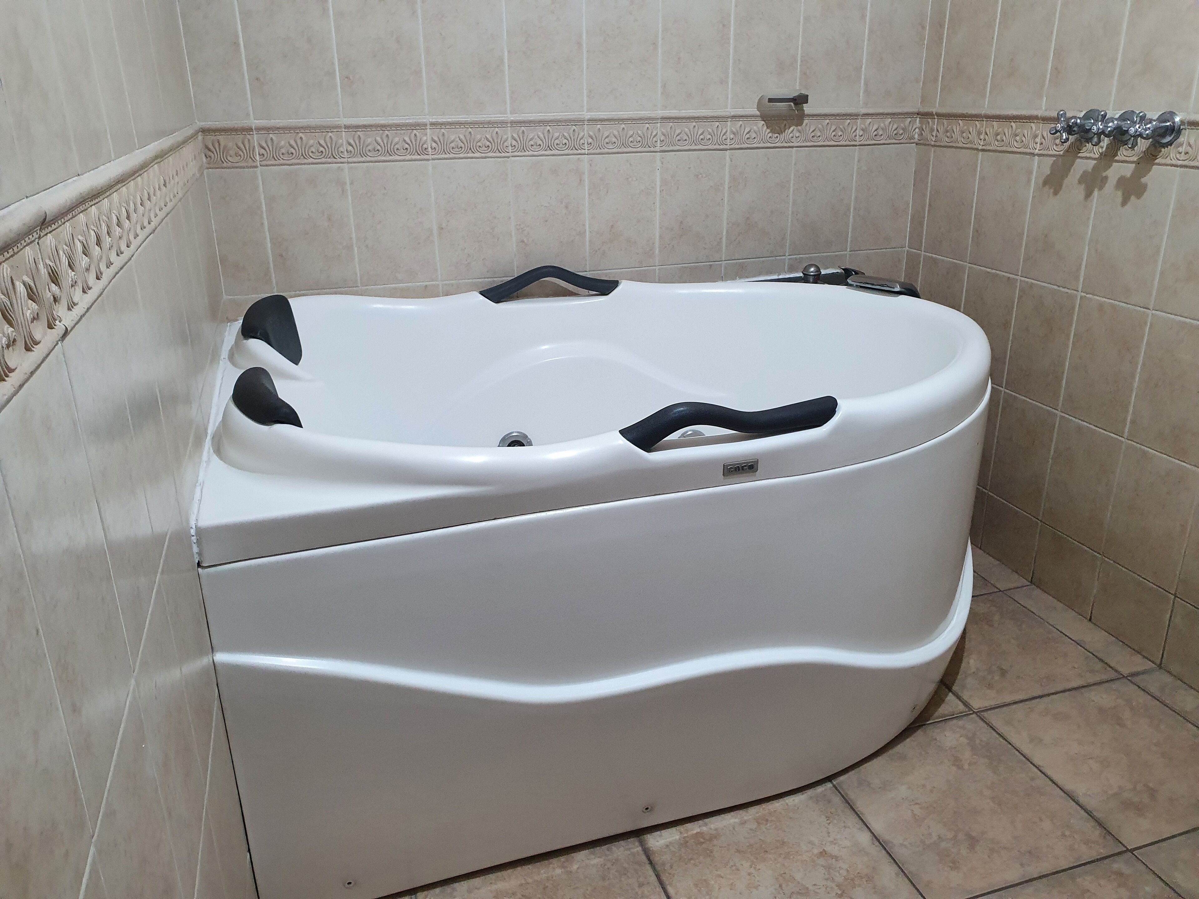 Een wit bad met een zwart handvat en bedieningspaneel, in een badkamer met beige betegelde muren en vloer.