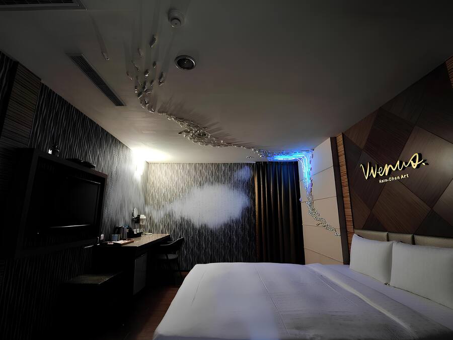 A modern hotel room with a large bed, a flat-screen TV, a desk with a chair, and a decorative wall with a logo.