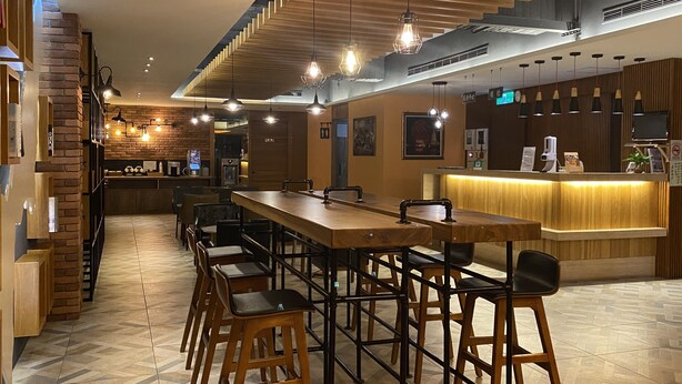 A modern restaurant with a long wooden bar, multiple seating options, and a counter with a digital menu display. - Huaxiang Hotel - Zuoying (Kaohsiung)