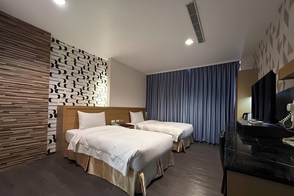 A hotel room with two beds, a wooden headboard, a patterned wall, a large window with curtains, and a desk with a telephone.