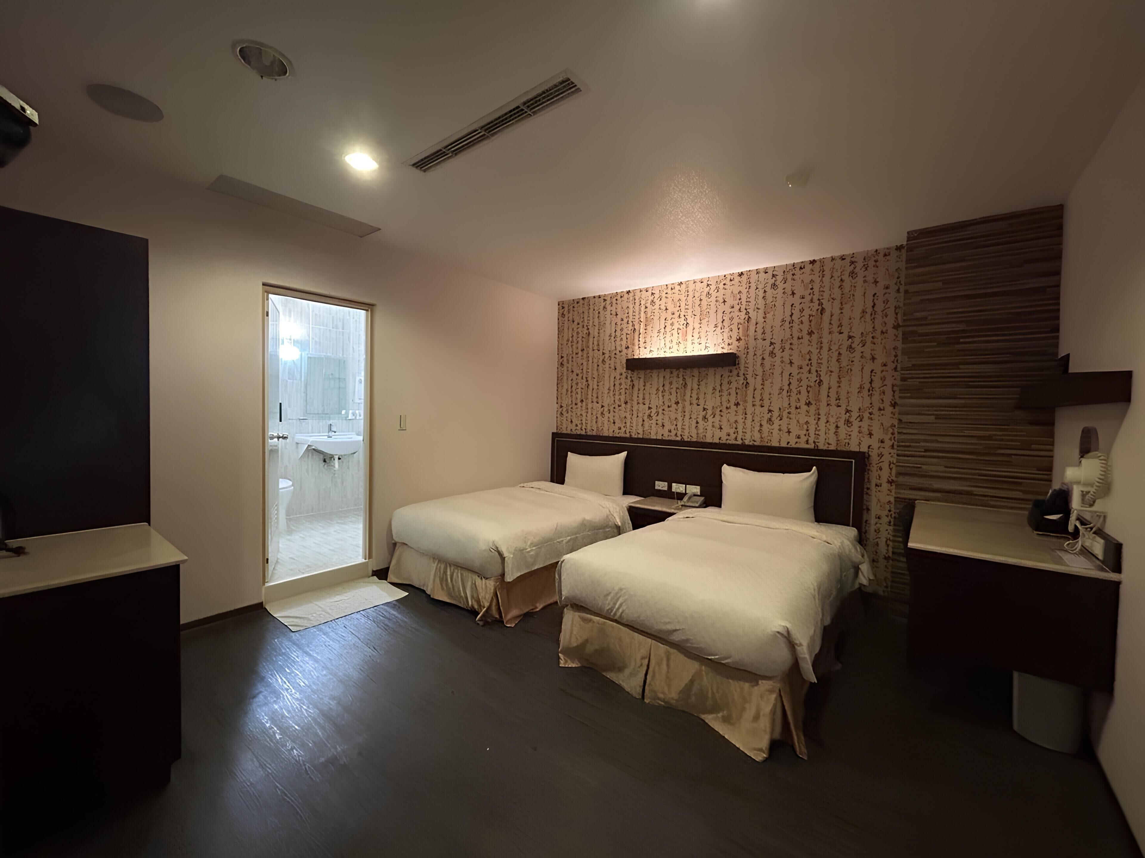 A hotel room with two beds, a bathroom with a sink and toilet, a small desk, and a TV.