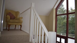 A staircase with a white railing, a yellow armchair with a striped pillow, and a large window with a view of trees.