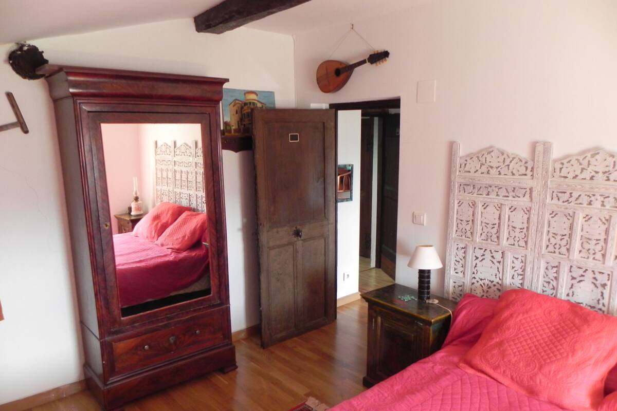 A bedroom with a wooden wardrobe, a bed with a red quilt, a small table with a lamp, and a guitar hanging on the wall.