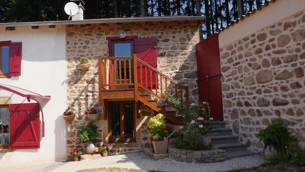A stone building with a wooden staircase leading to a balcony, surrounded by potted plants and flowers. - Ferme des Entremains (Thizy-les-Bourgs)
