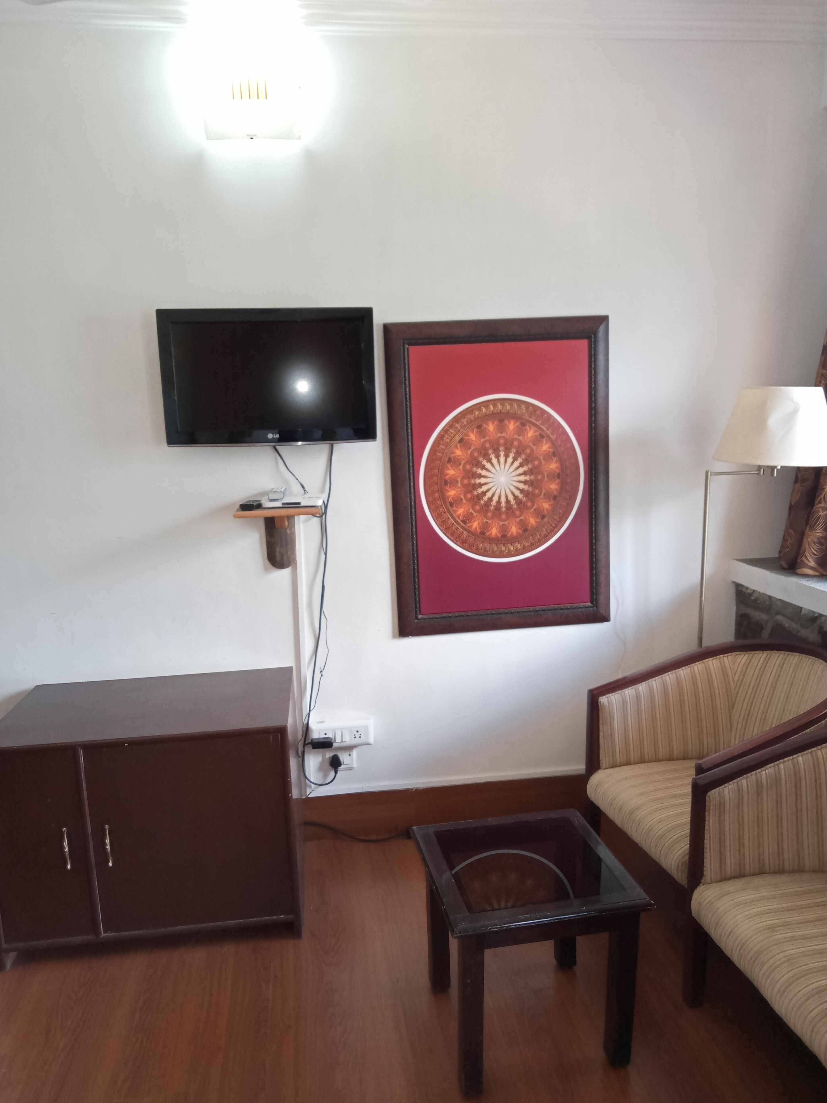 Un salon avec un téléviseur à écran plat fixé au mur, une œuvre d’art représentant un mandala encadré, un meuble en bois foncé, une table basse en verre, un canapé à rayures et un lampadaire.