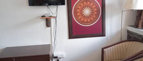 A living room with a flat-screen TV mounted on the wall, a framed mandala artwork, a dark wooden cabinet, a glass coffee table, a striped sofa, and a floor lamp.