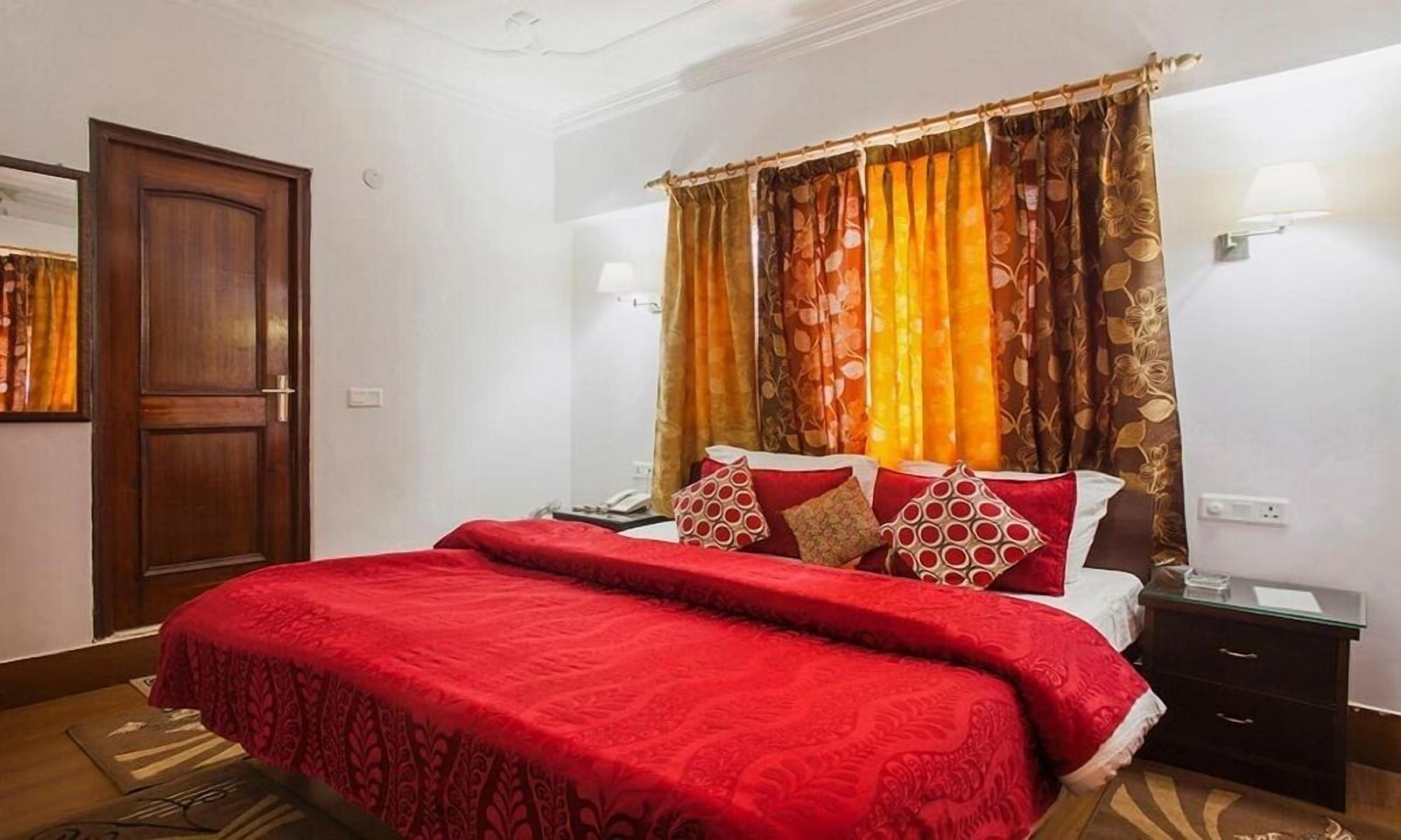 A hotel room with a large bed, red bedding, a wooden door, a nightstand, and a mirror.