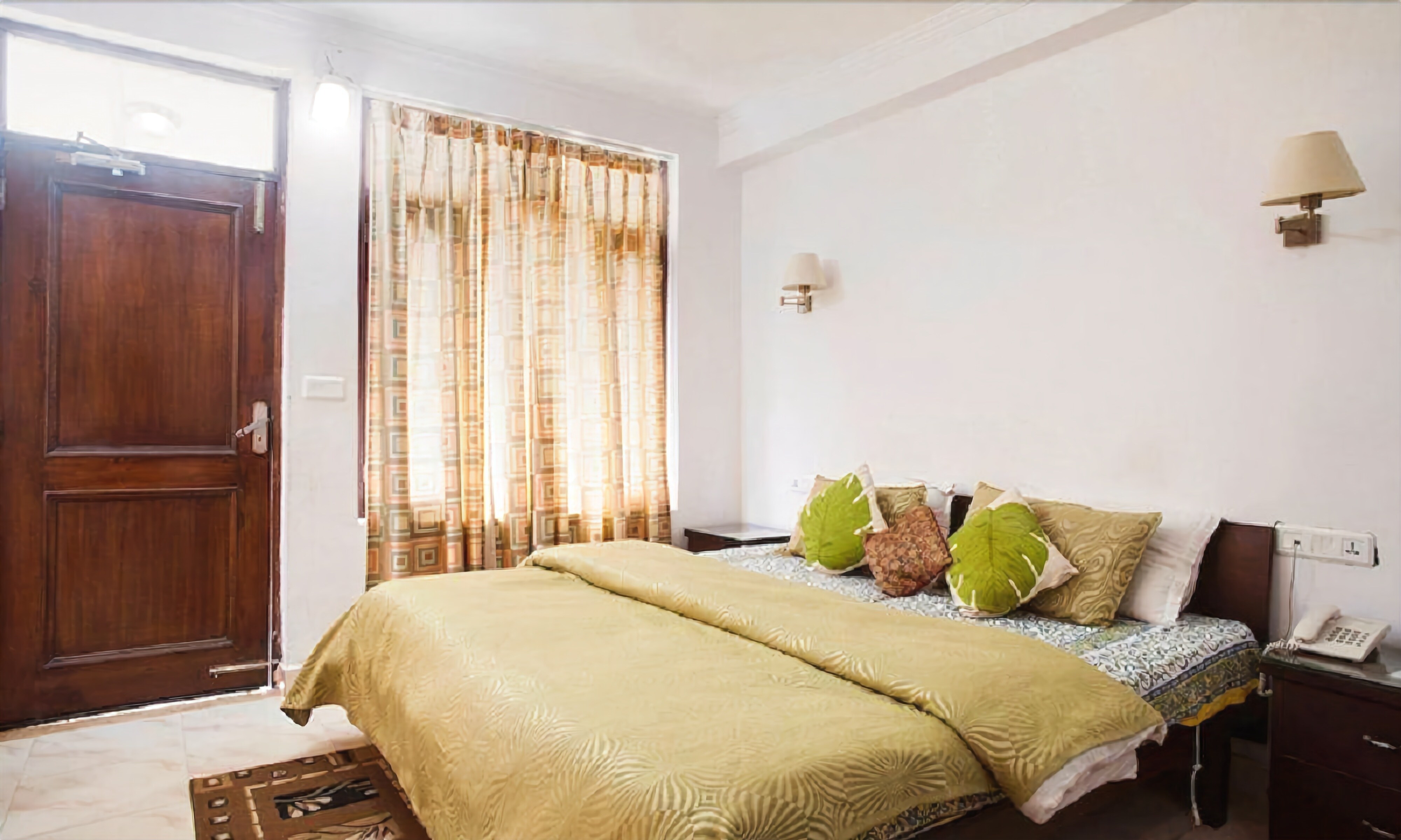 A bedroom with a wooden door, patterned curtains, a bed with multiple pillows, a bedside lamp, and a telephone on the nightstand.