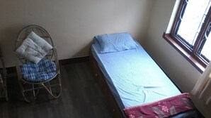 Una camera piccola con un letto singolo, pavimento in legno, una sedia in rattan con cuscini e una finestra che lascia entrare luce naturale.