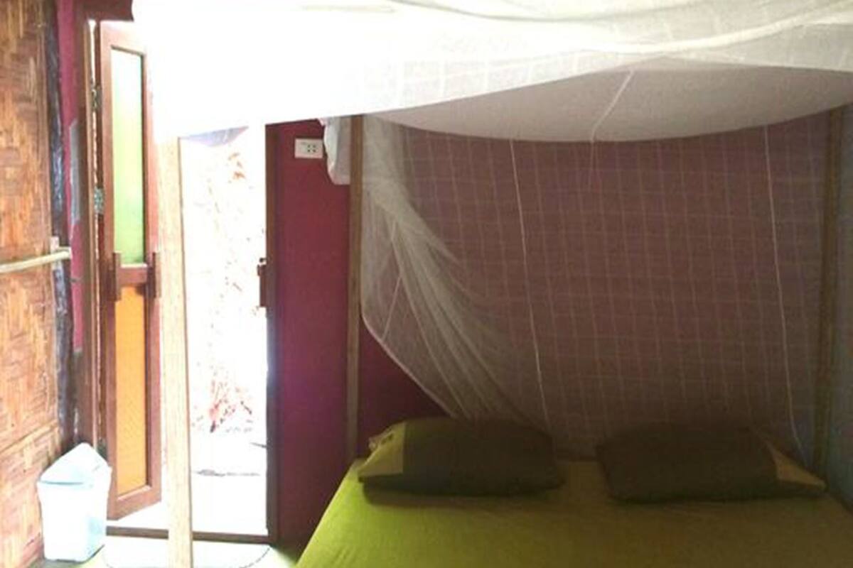 Una cama individual con mosquitera, almohadas verdes y rojas, y piso de madera.