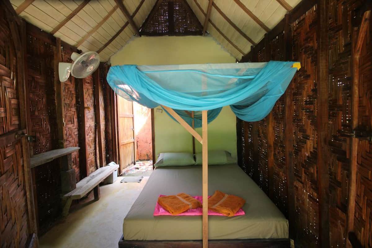Una cama individual con mosquitero, un banco de madera y un ventilador en una habitación rústica con paredes y techo inclinados de madera.