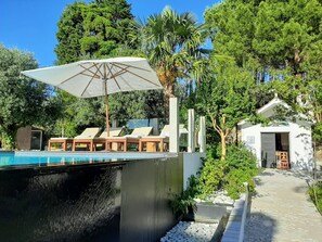 Ein Pool mit Liegestühlen und Sonnenschirm, umgeben von Grünpflanzen und einem Gebäude mit einer Tür.