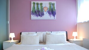 A neatly made bed with white linens and pillows, a heart-shaped decoration with lavender, and two bedside lamps on a purple wall.