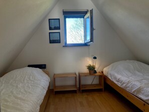 A small, triangular attic bedroom with two single beds, a wooden nightstand with a lamp, and a window with a view of trees.