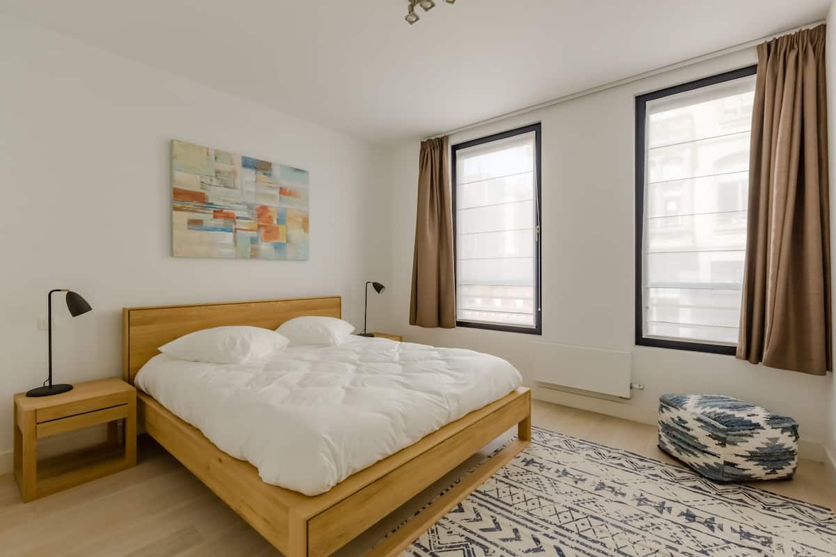 Un dormitorio con cama de madera, mesita de noche, lámpara de pie, alfombra estampada y ventana con cortinas.