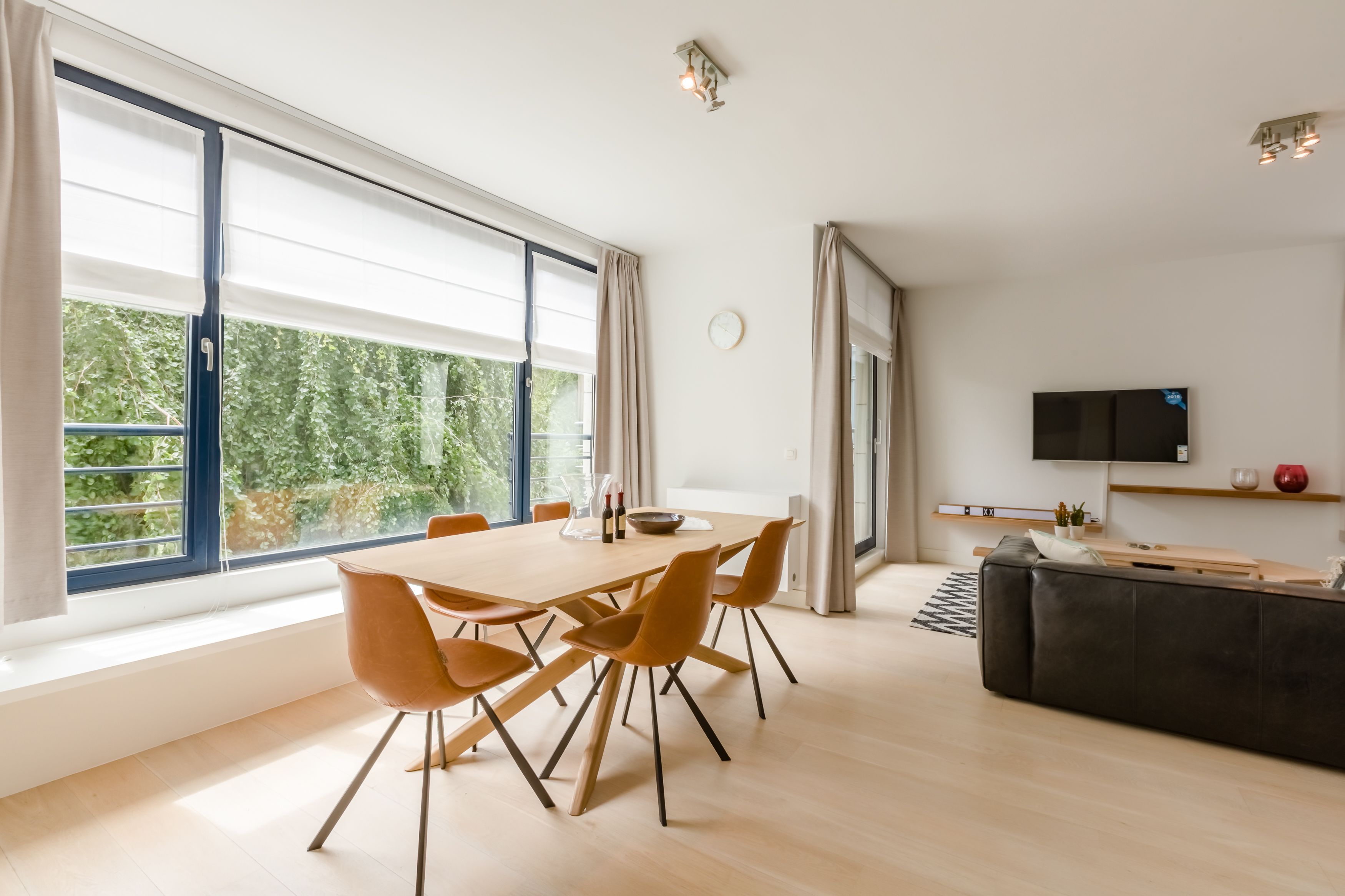 A modern living room with a dining area, large windows, a flat-screen TV, and a black leather sofa.