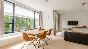 A modern living room with a dining area, large windows, a flat-screen TV, and a black leather sofa.