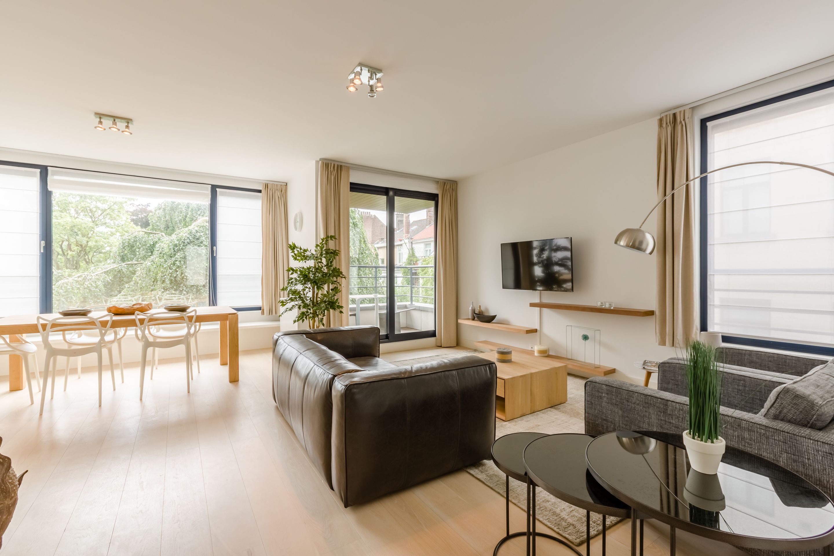 Een moderne woonkamer met een bank, eettafel en stoelen, een televisie, en grote ramen met gordijnen.
