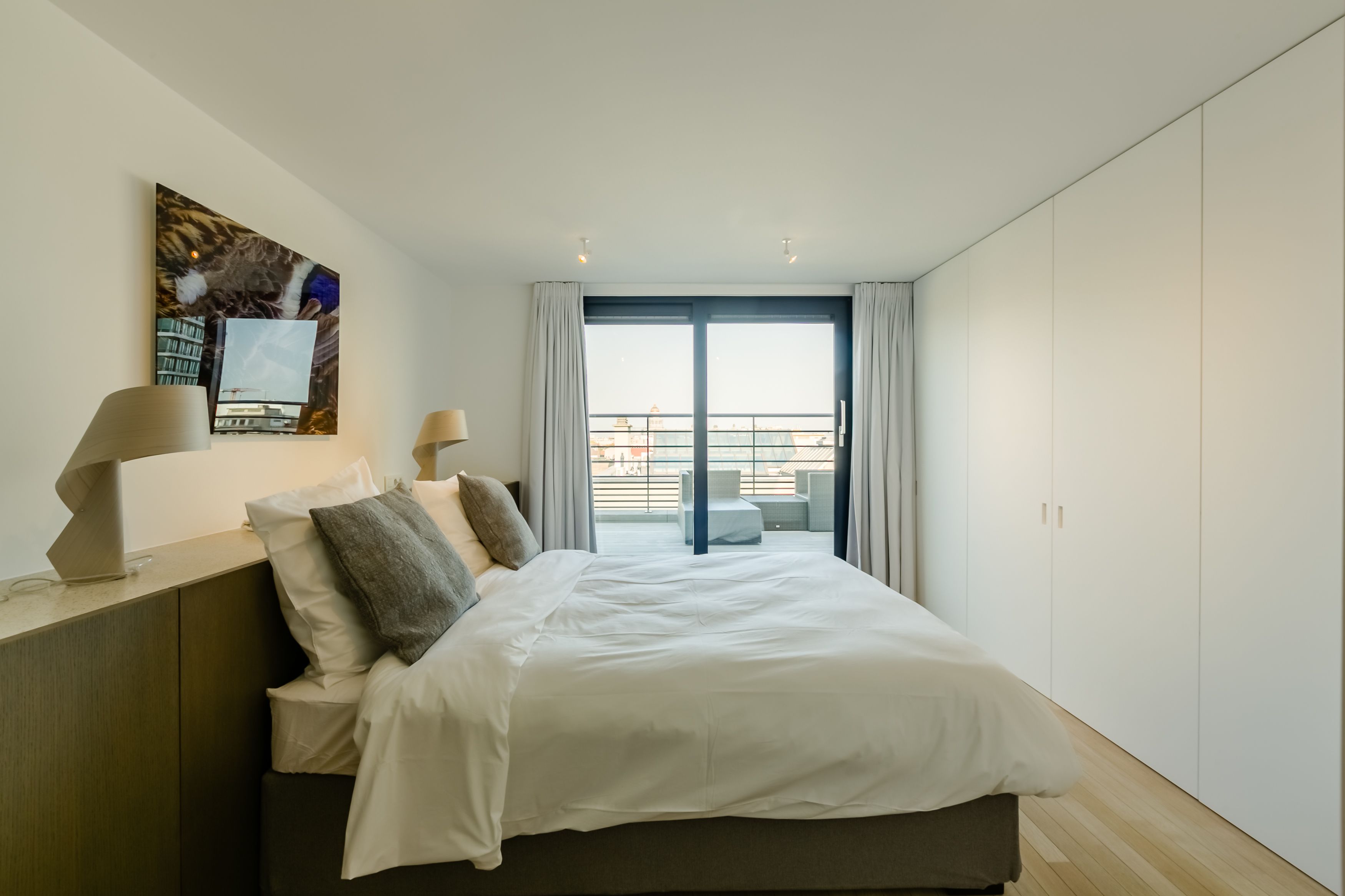 Moderni hotellihuone, jossa on suuri sänky, työpöytä lampun kanssa, taulu seinällä ja näkymä kaupunkiin parvekeovien kautta.