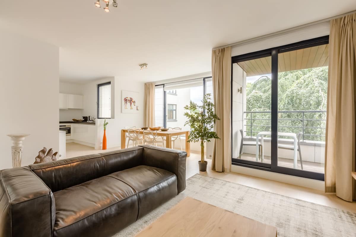 Una sala de estar moderna con un sofá de cuero, un comedor con mesa y sillas, y un amplio ventanal corredero que da a un balcón con mobiliario exterior.
