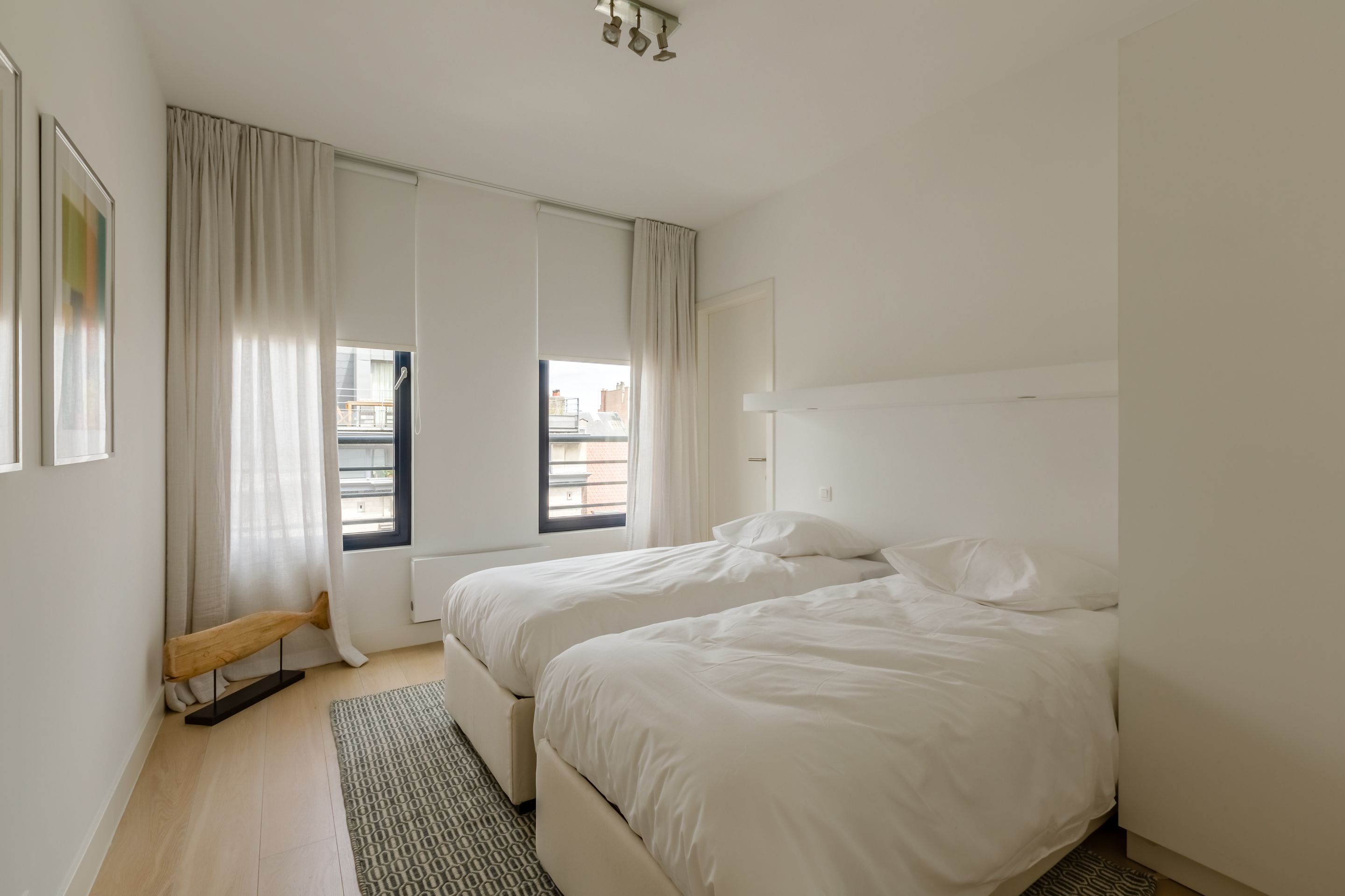 A modern hotel room with two beds, a large window with curtains, a radiator, and a patterned rug.