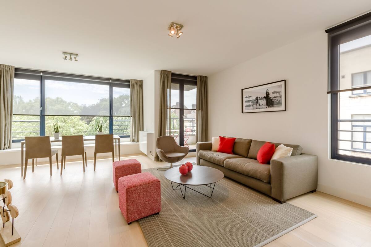 Una sala de estar moderna con sofá, reposapiés rojo, mesa de centro redonda y una silla. Amplios ventanales ofrecen vistas al exterior, con vegetación.