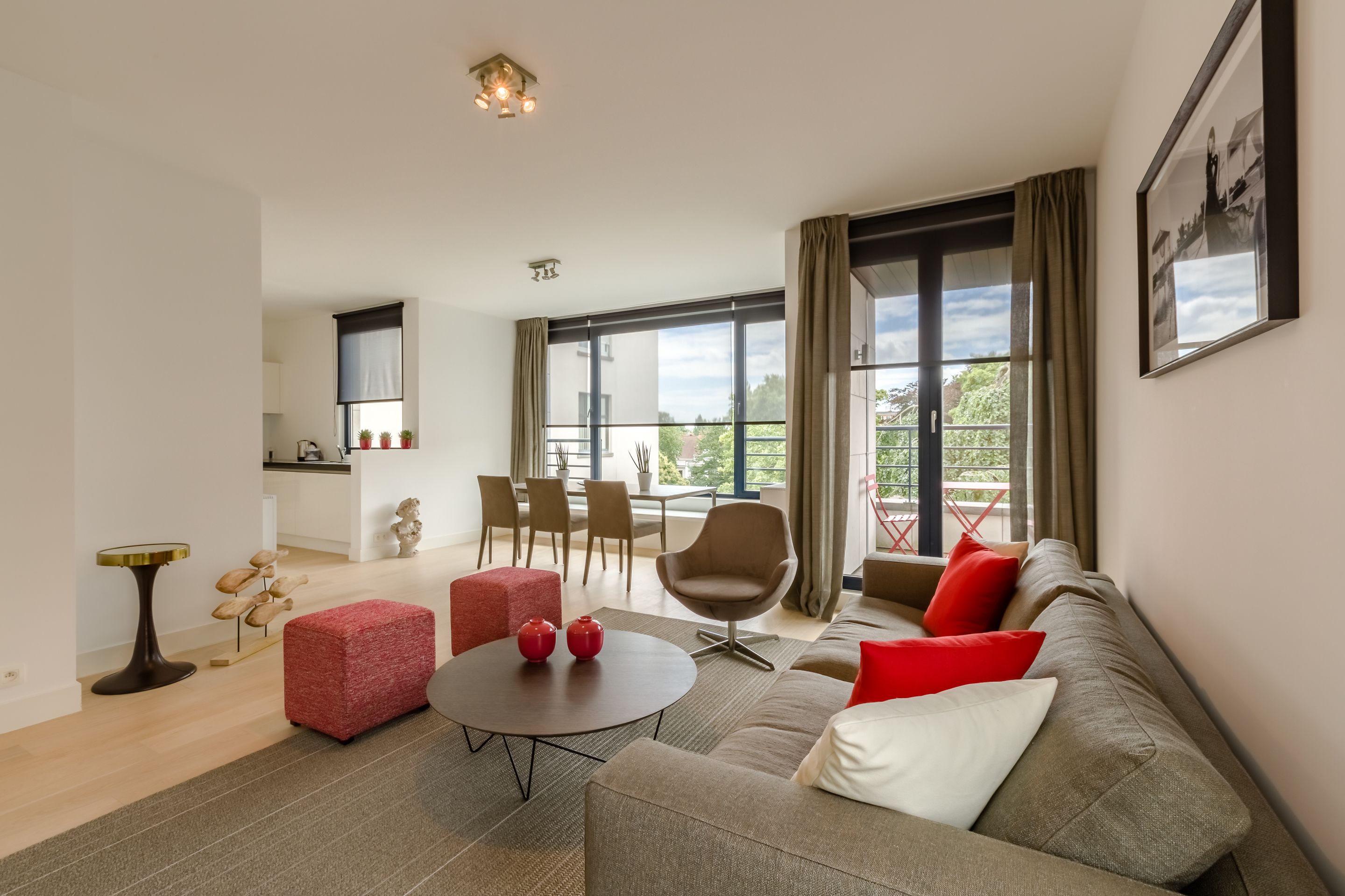 Een moderne woonkamer met een bank, een rode poef, een ronde salontafel en een eethoek met stoelen en een tafel.