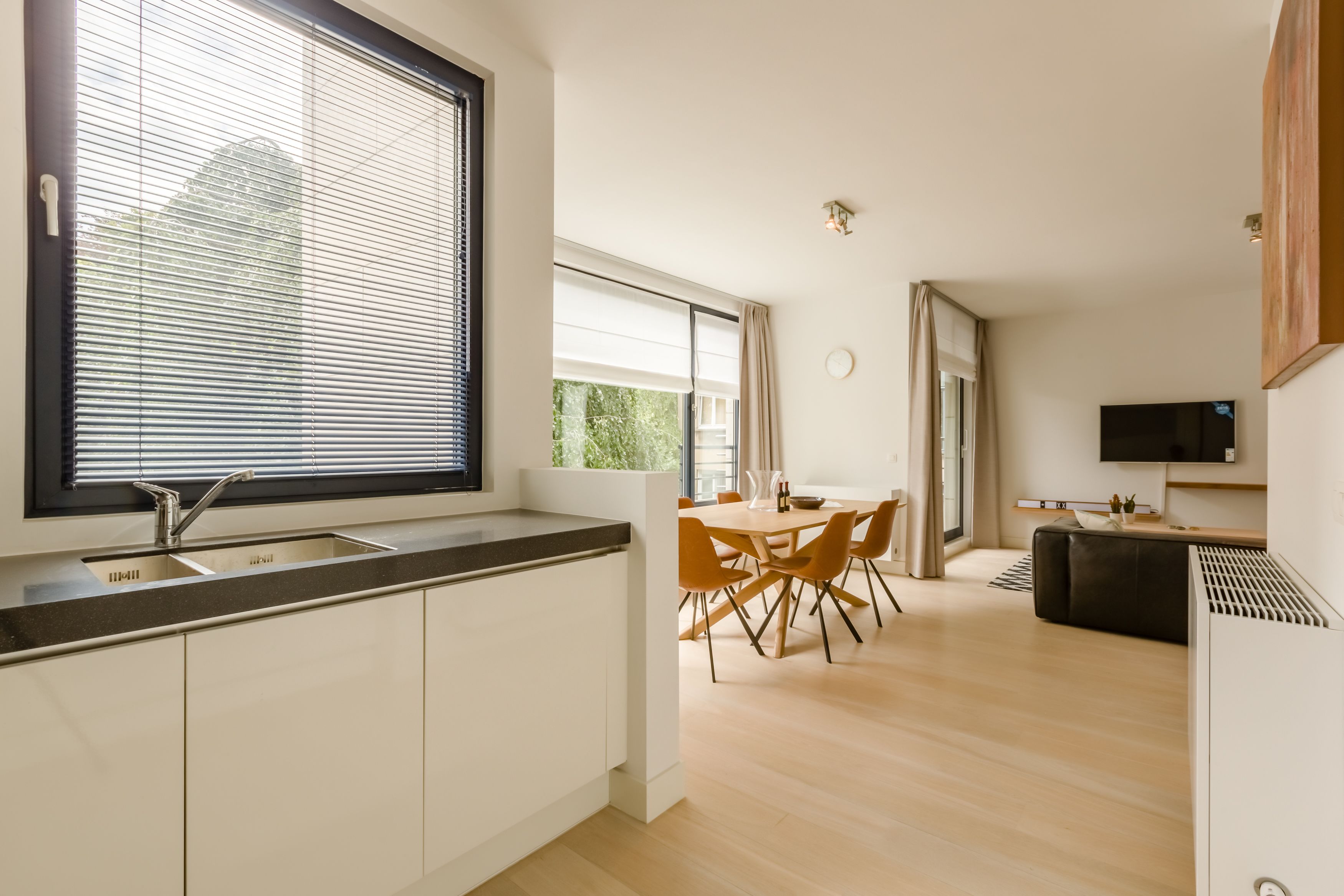 Cozinha moderna com uma janela grande, área de refeições com mesa e cadeiras, e sala de estar com sofá e televisão.