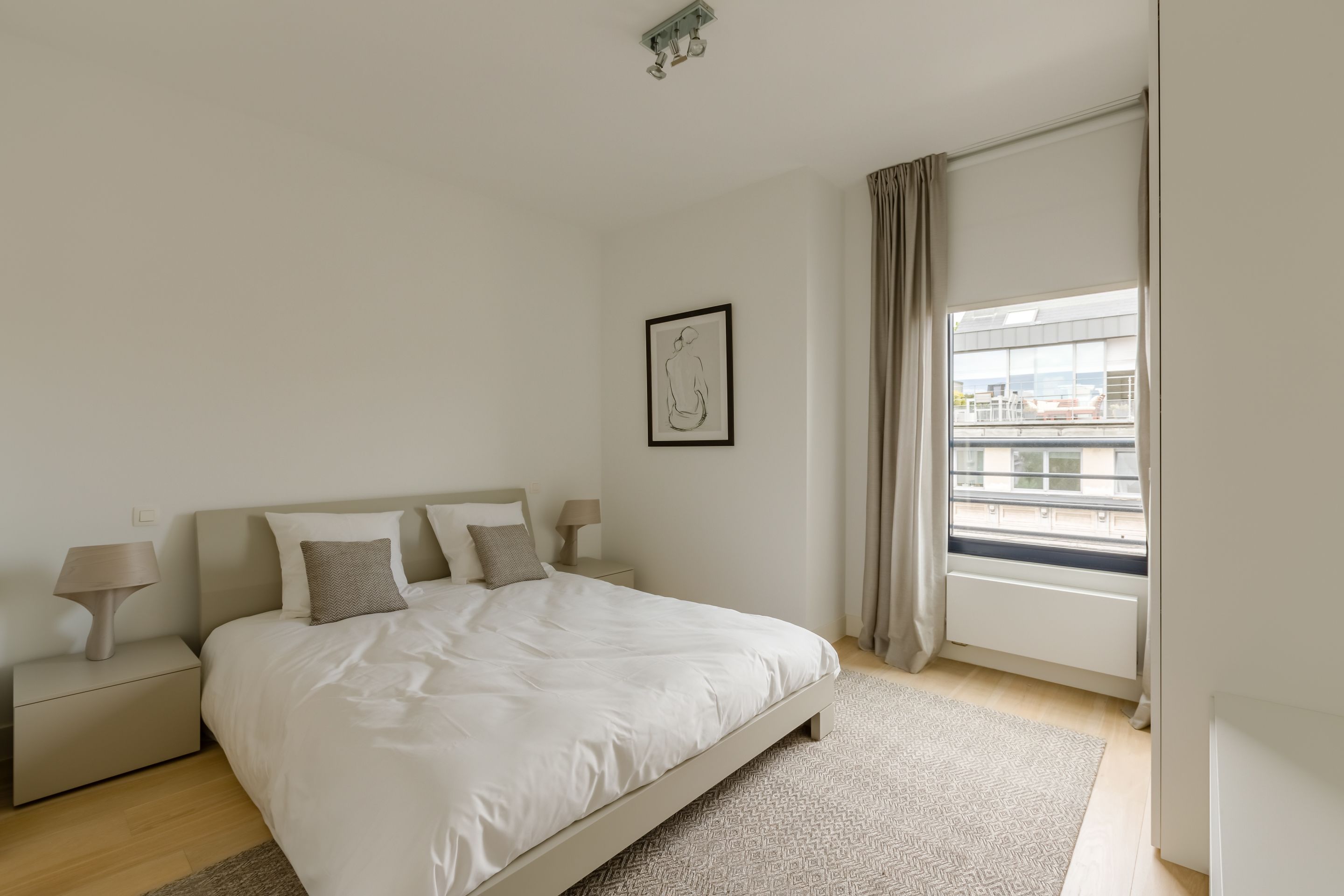A modern bedroom with a large bed, a nightstand, a lamp, a framed picture on the wall, and a window with curtains.