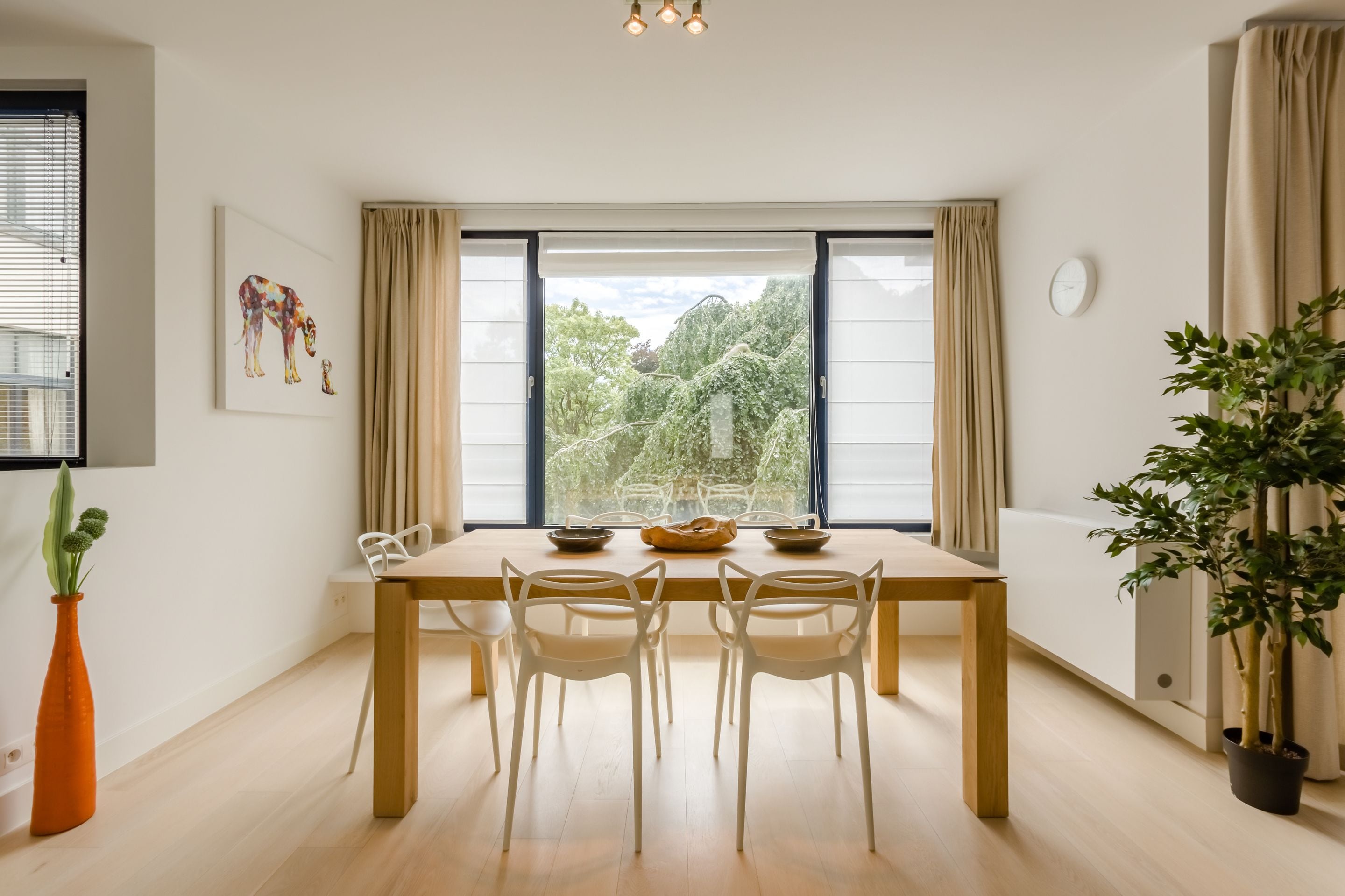 A dining area with a wooden table and white chairs, a vase with green plants, and a window with curtains.