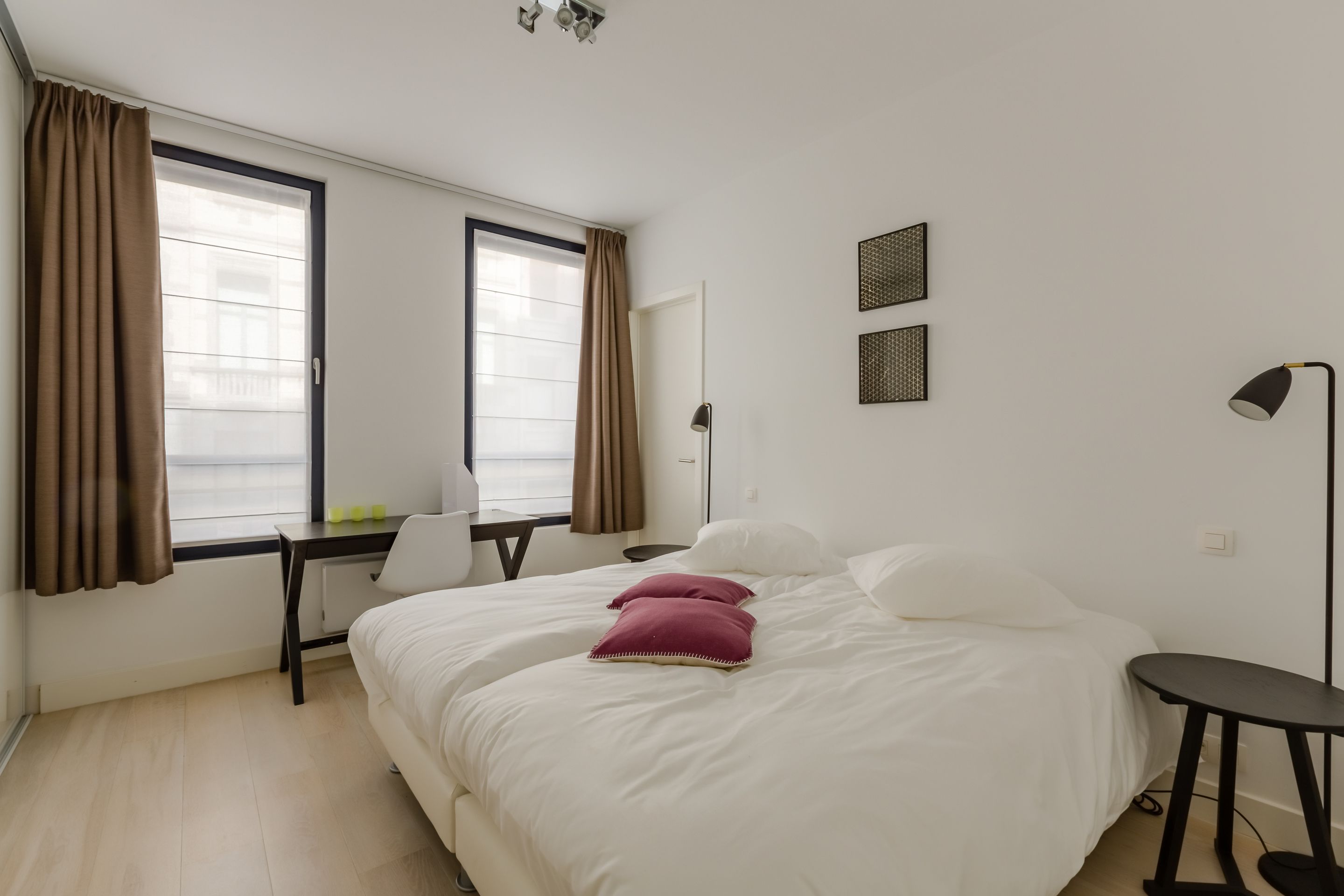 A hotel room with a large bed, a desk, and a chair. There are two windows with curtains, a lamp, and a wall-mounted shelf.