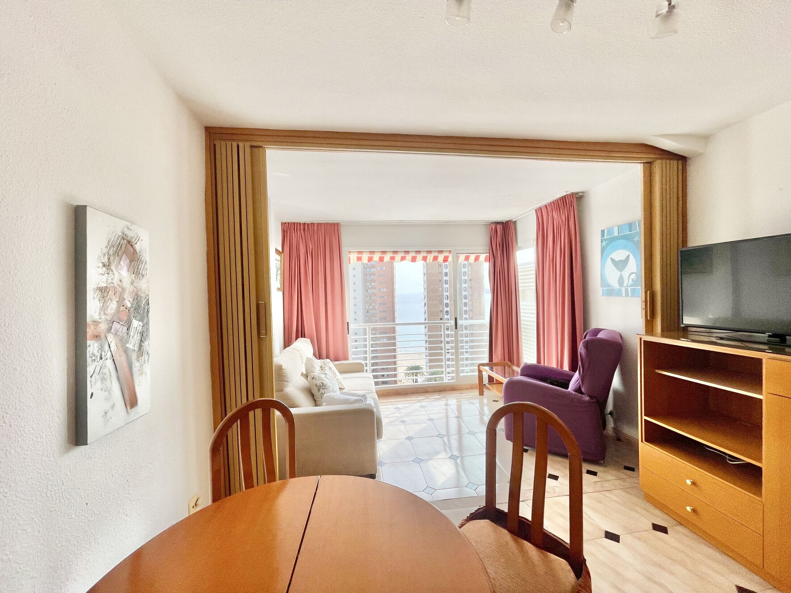 A living room with a dining table, chairs, a sofa, a television, and a balcony with curtains.