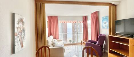 A living room with a dining table, chairs, a sofa, a television, and a balcony with curtains.