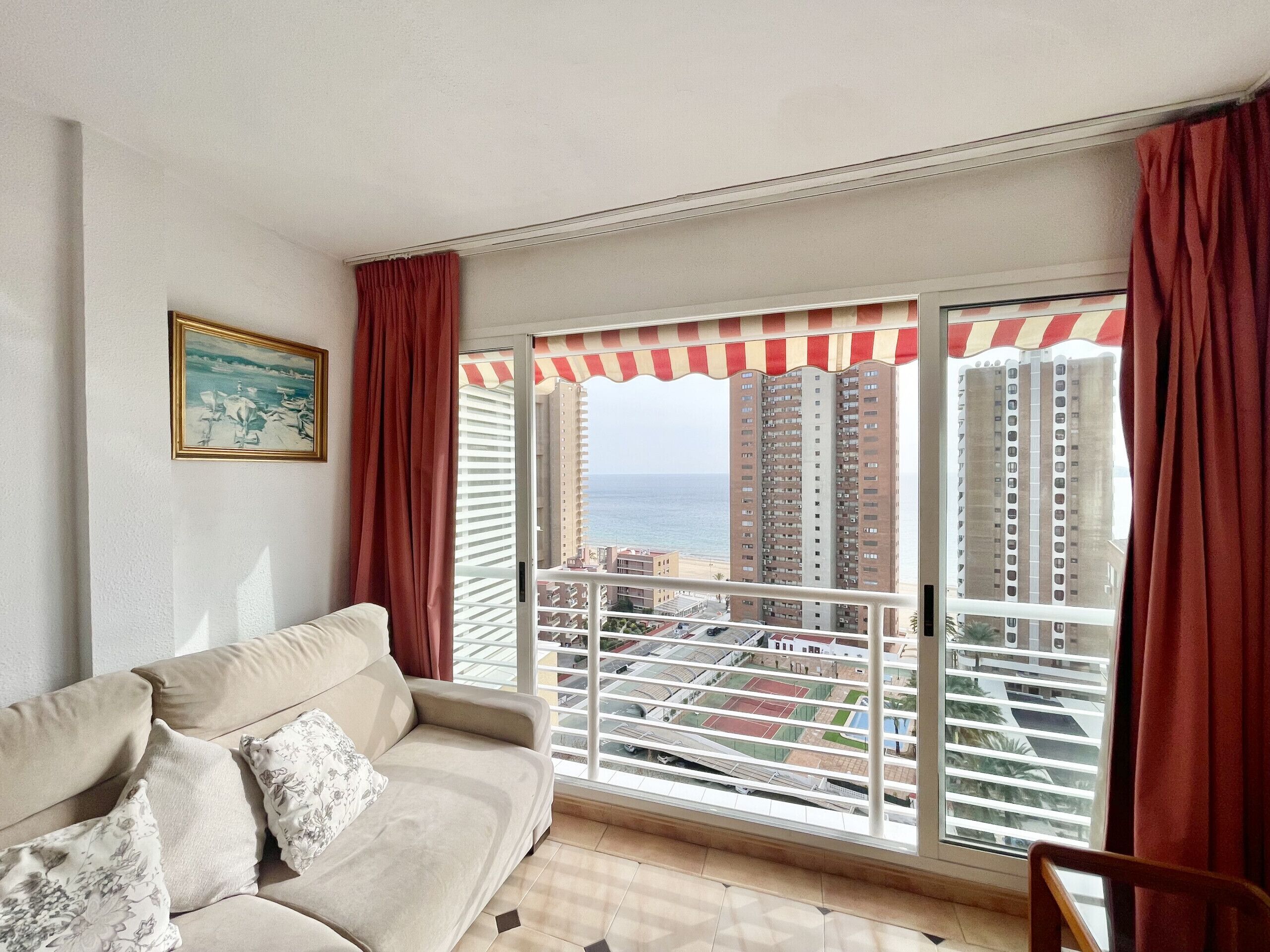 A living room with a beige sofa, red curtains, and a view of tall buildings and the sea through a balcony with a striped awning.