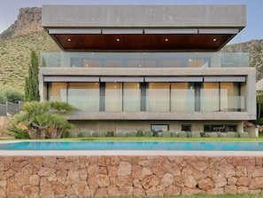 A modern two-story house with a stone facade, large windows, and a swimming pool in the foreground.