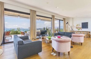 A spacious living room with a large window offering a view of the sea and mountains. There are two sofas and a coffee table, with a dining area visible in the background.