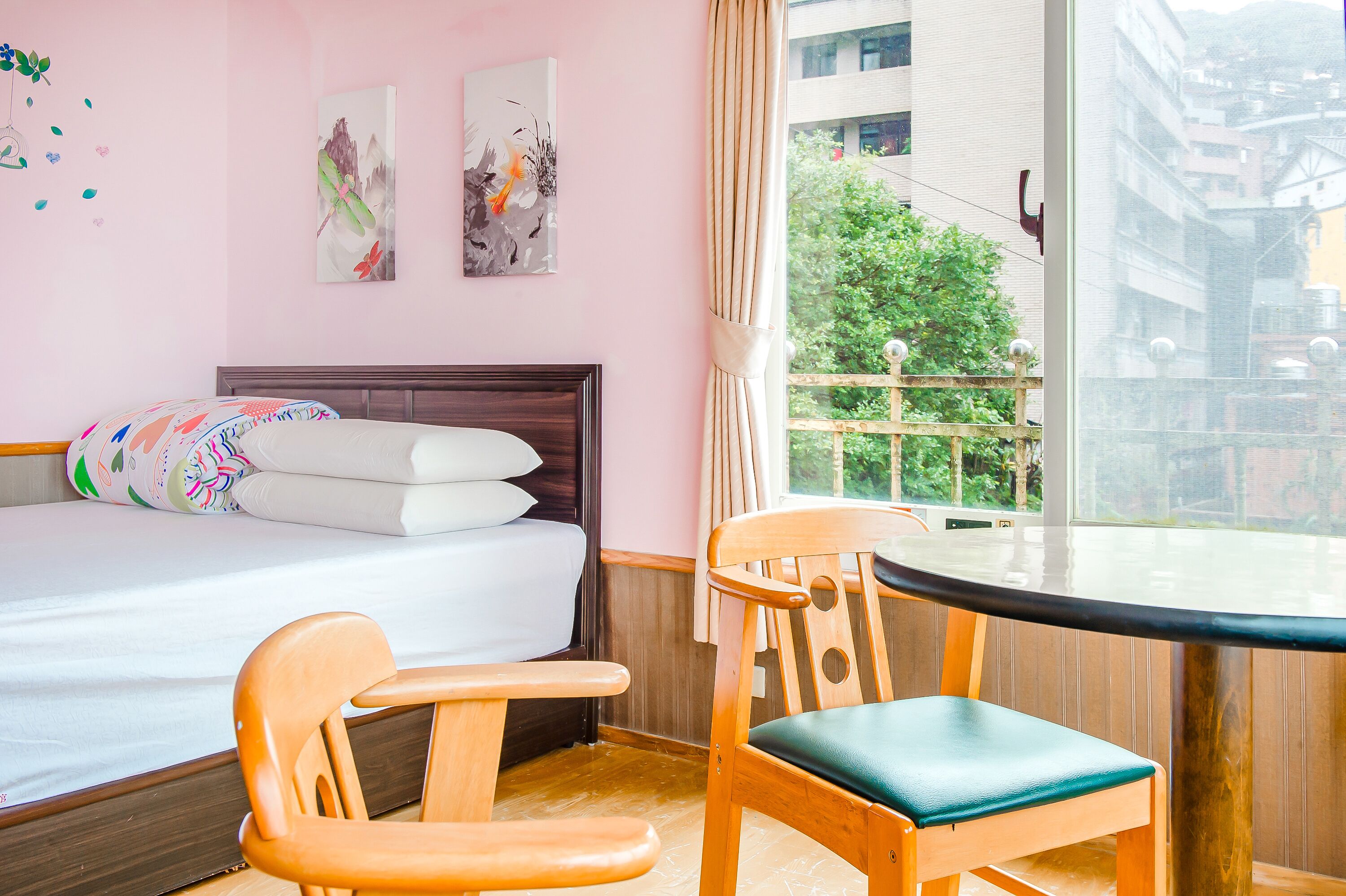 A hotel room with a bed, a wooden chair, a small table, and a window with a view of buildings and trees.