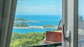 A coastal view from a window overlooking a cityscape with a red brick building and lush greenery. - CF Kite Homestay (New Taipei City)
