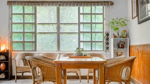 A dining area with wicker chairs and a wooden table, large windows with a view of trees, a shelf with decorative items, and a wall-mounted shelf with plants. - CF Kite Homestay (New Taipei City)