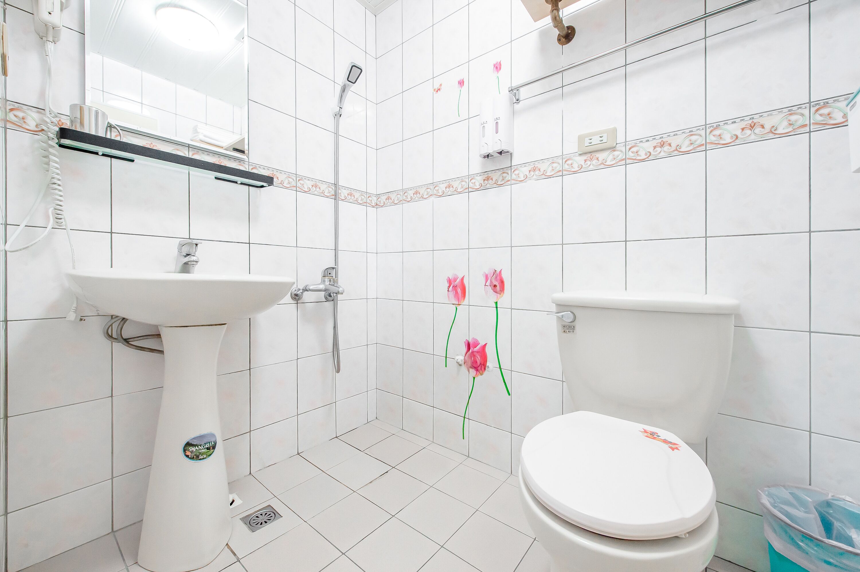 Une salle de bain avec un lavabo, des toilettes et une douche, le tout de couleur blanche. Les murs sont décorés de carreaux et un petit étagère se trouve au-dessus du lavabo.